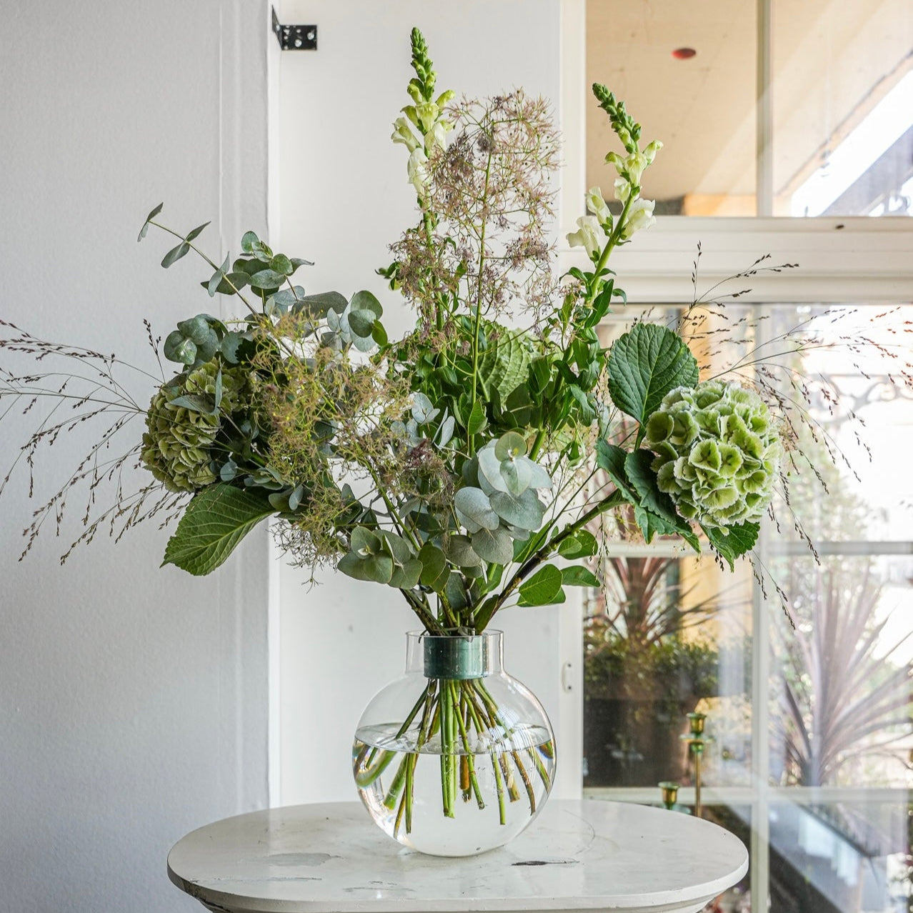 A clear glass vase with an arrangement of green and white flowers, including hydrangeas and eucalyptus, sits on a small white round table. The Hanataba Ivy Green bouquet stands out as sunlight streams through a tall window in the background.
