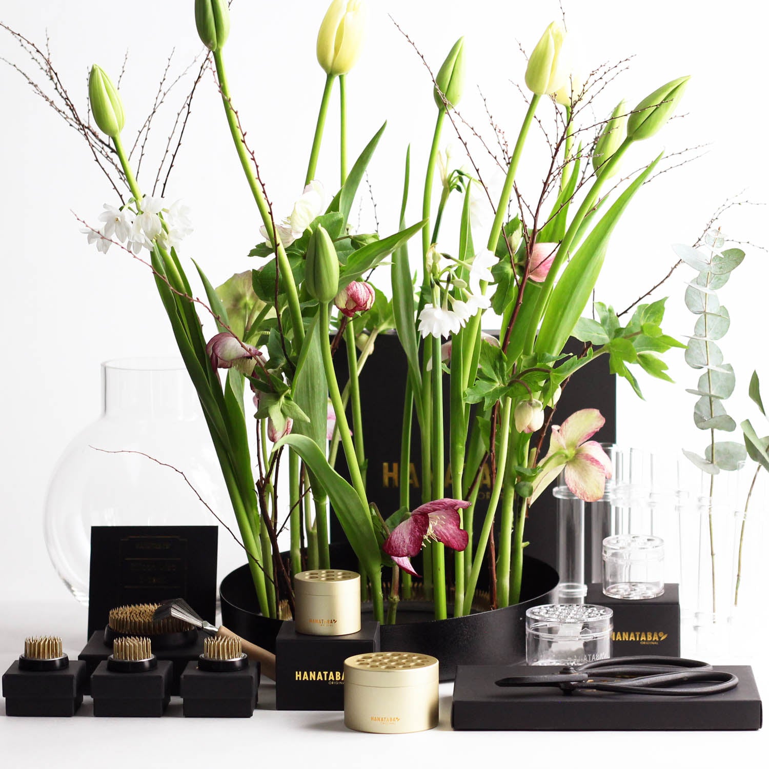 Flowers arranged in a black tray surrounded by a rich variety of flower arrangement tools, flower frogs and vases