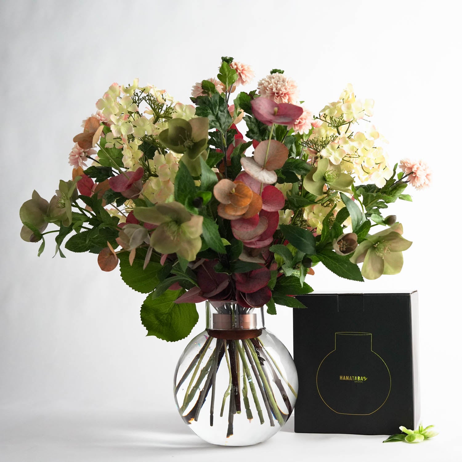 Bouquet of fresh pink and white flowers arranged in a globe shaped glass vase filled with water. The "Hanataba Bubble Glass Vase" also contains a "Hanataba Bouquet Twister" in the pink champagne color. The glass vase is placed in a white background next to a black packaging box with the Hanataba logo printed in gold.