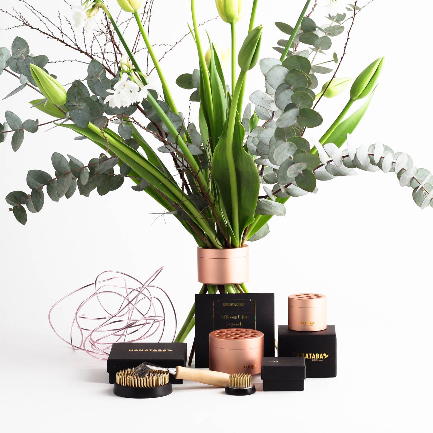 The Home Florist’s Starter Kit – Pink Champagne by Hanataba features an elegant bouquet in a vase with eucalyptus leaves and tulips, surrounded by twisted pink bonsai aluminum wire. The kit includes Hanataba Flower Twisters in pink champagne, a 70 mm flower frog, a 34 mm flower frog, a kenzan brush, and black Hanataba packaging boxes, all beautifully arranged on a white surface.