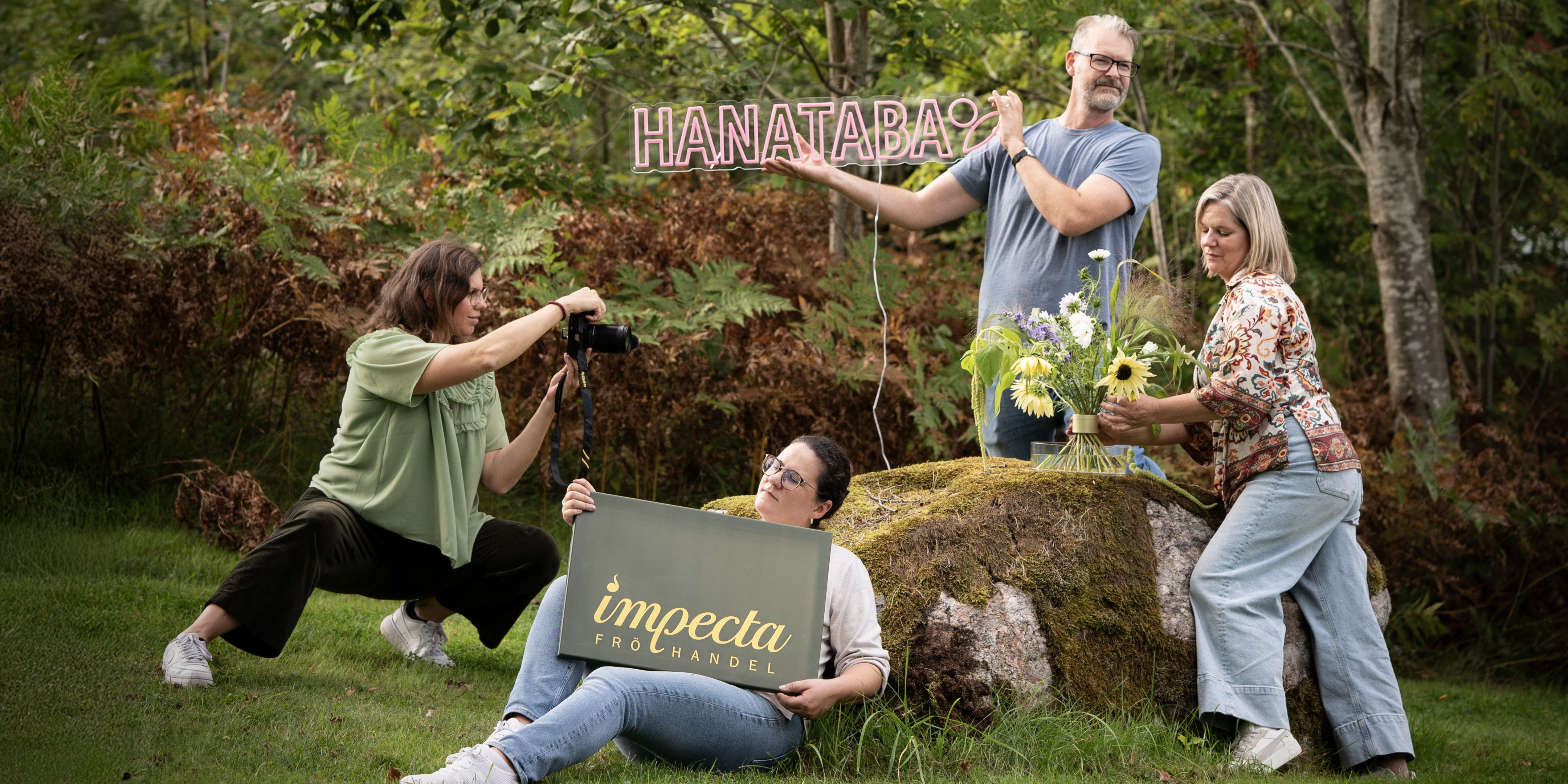 Group of people outdoors with 'HANATABA' and 'impecta' signs in a natural setting.
