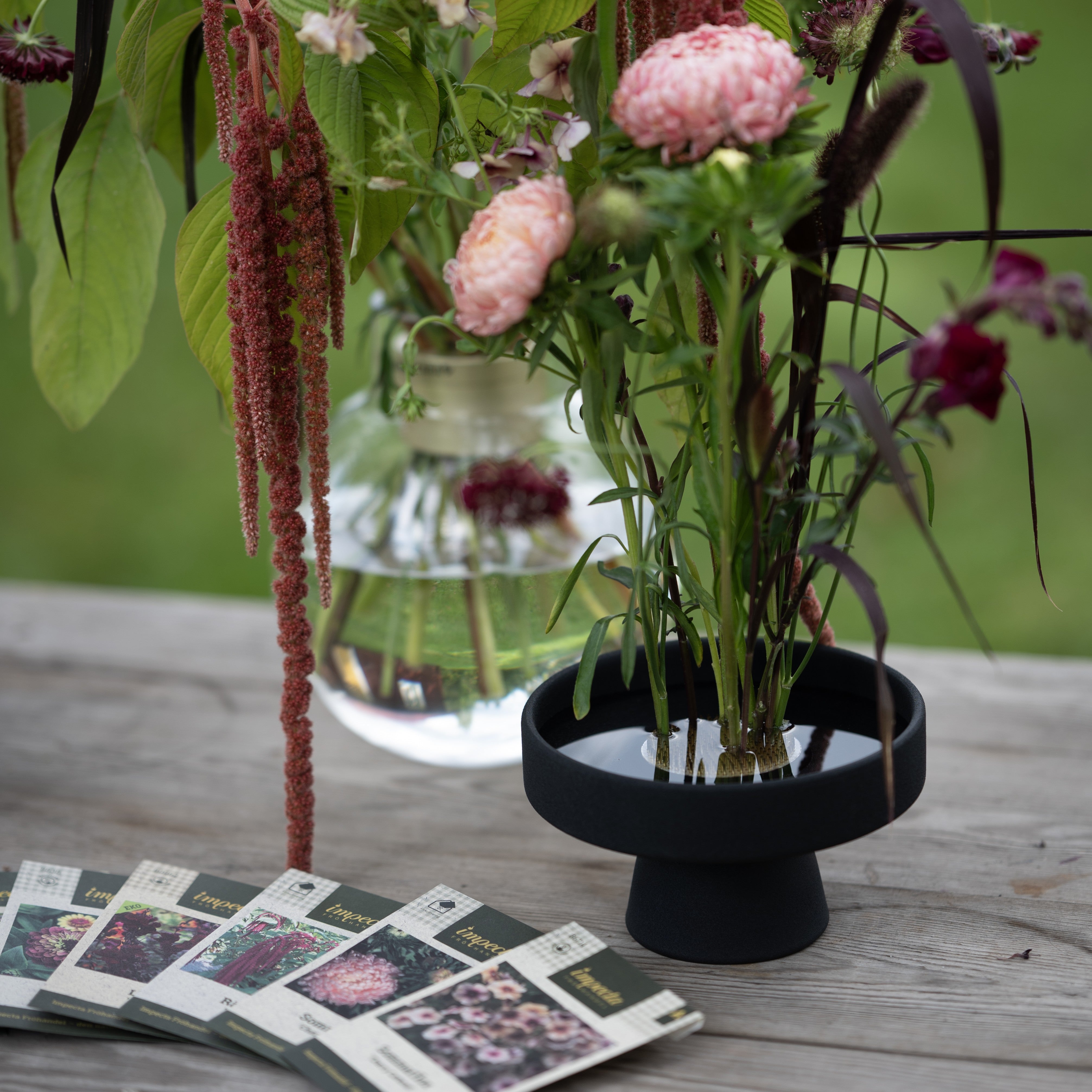 Floral arrangement in a black hanataba ceramic ikebana vase on a wooden surface with seed packets from impecta fröhandel.