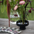 Floral arrangement in a black hanataba ceramic ikebana vase on a wooden surface with seed packets from impecta fröhandel.