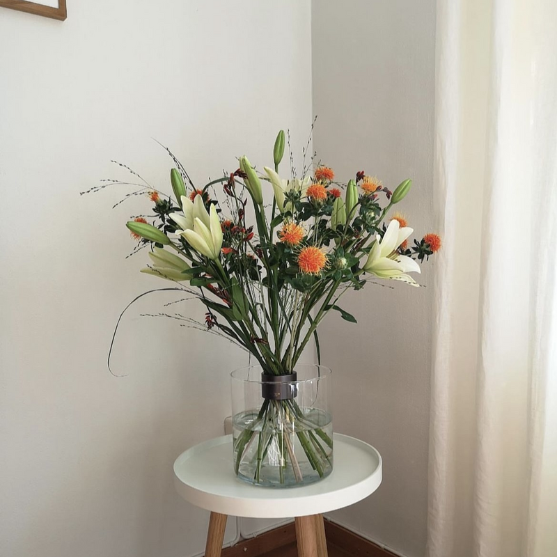 Minimalist Ikebana-style floral arrangement featuring white lilies and orange Carthamus in a clear vase with a Hanataba Spiral Stem Holder on a white table.