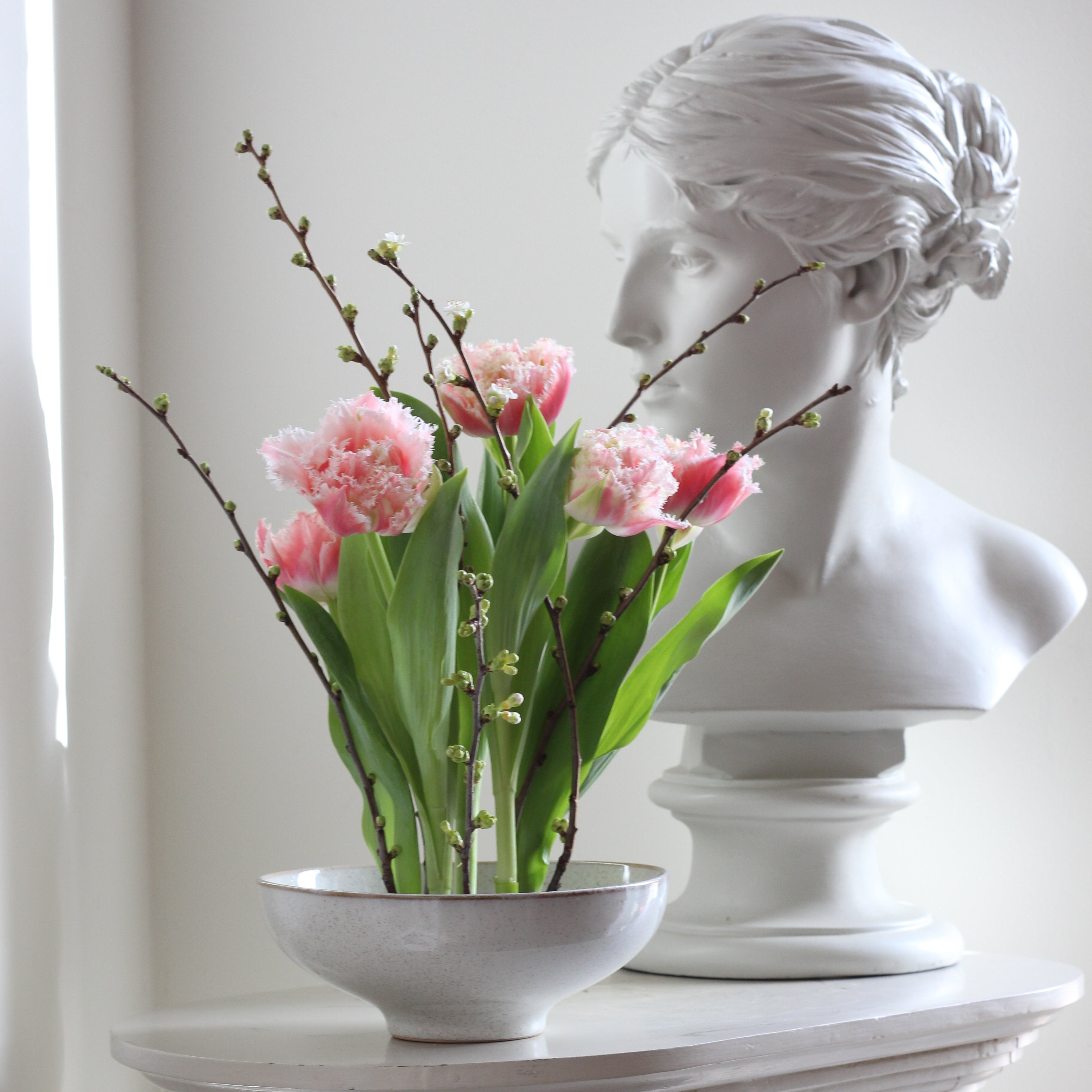 Arrangement of pink tulips and budding branches anchored by a 120mm Kenzan Fakir medium ring, set against a classical white bust sculpture and grey wall.