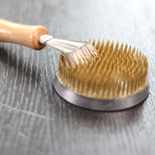 A kenzan rake with a wooden handle being used to comb through the brass needles of a golden kenzan, set on a wood-toned surface.
