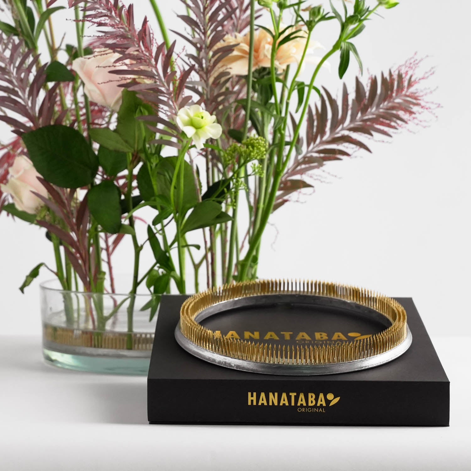 A kenzan flower frog ring in a glass vase with a lush bouquet of pastel flowers and green foliage. In front of the flowers a black packaging box with "HANATABA ORIGINAL" in gold letters with another flower frog on top of it. Behind it, the scene is set on a white surface with a plain background.