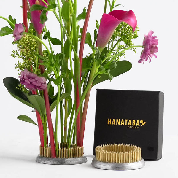 A flower frog bouquet of pink and purple flowers in a 70 mm kenzan ring with golden spikes. Next to it is a black box labeled "Hanataba Original." The setup is on a white background.