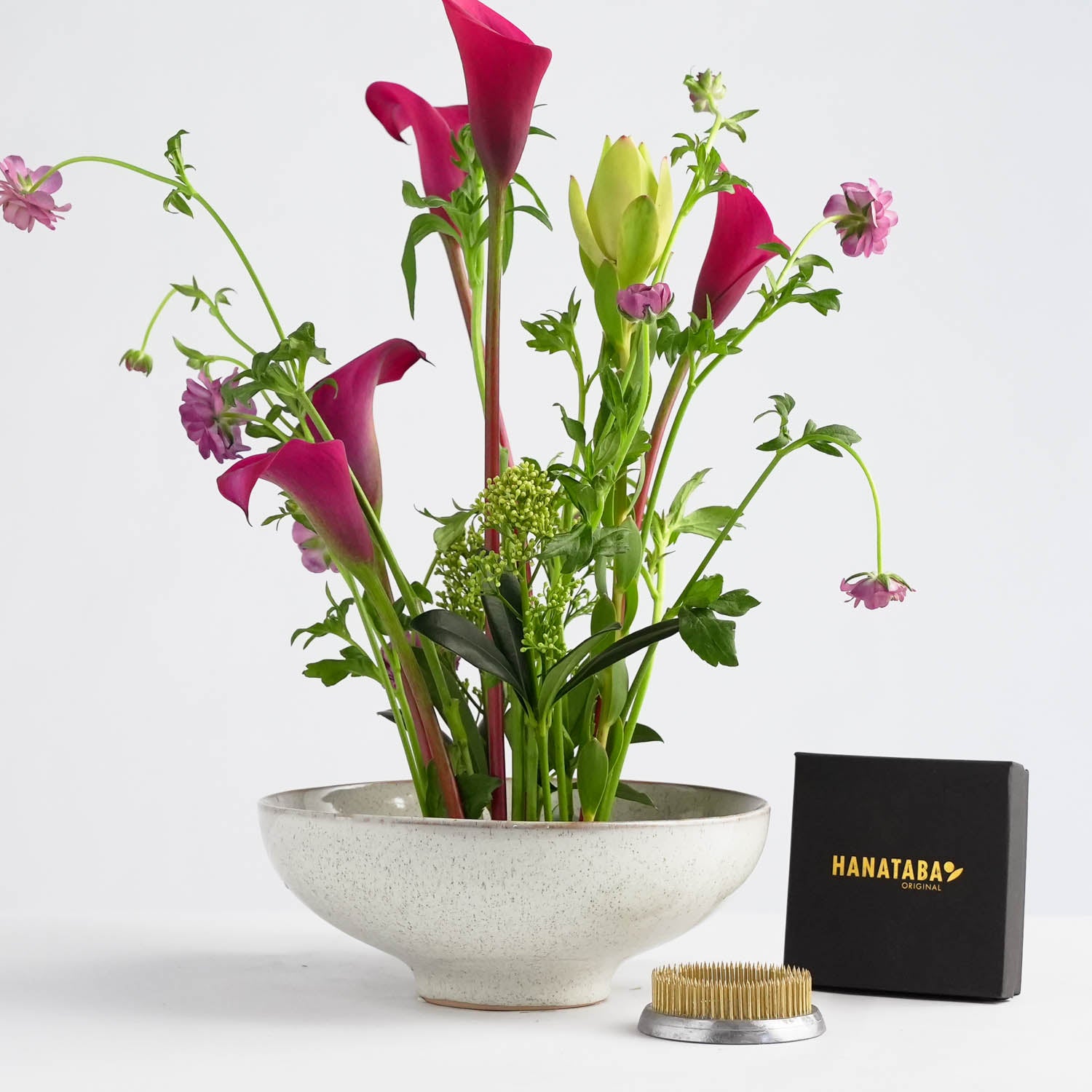 An intricate 70 mm kenzan positioned at the base of a floral arrangement, with supporting lush green stems with red fresh flowers, set against a textured ceramic dish.