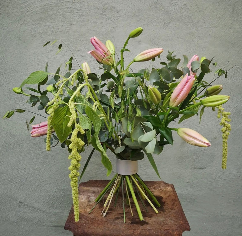 A lush floral arrangement featuring pink lilies and cascading green accents, all held together by a Hanataba Bouquet Twister in pearly silver, against a textured grey wall