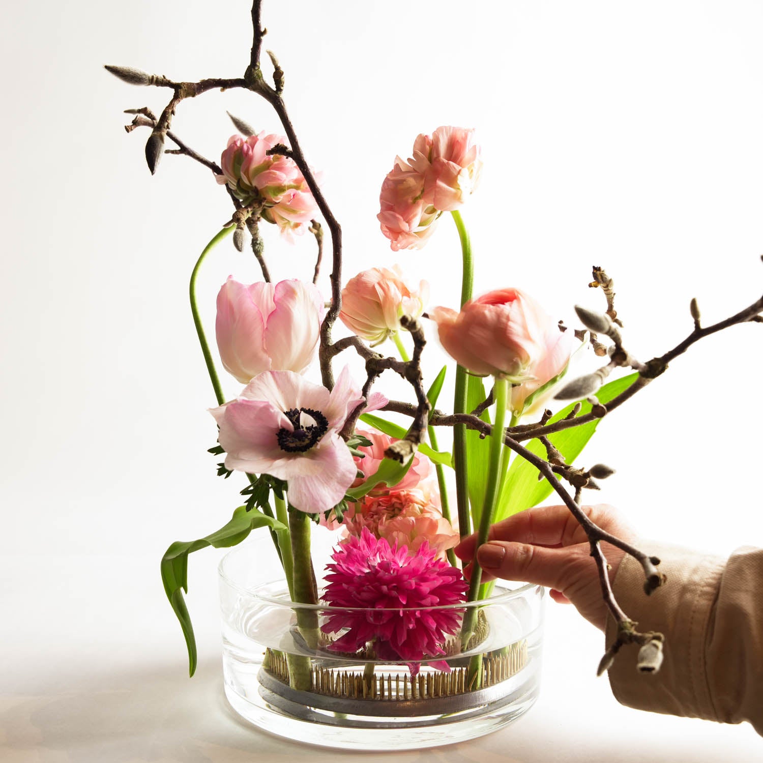 Florist arranging pink and purple flowers in a clear glass bowl filled with water. The flower arrangement is set in a bright white setting.