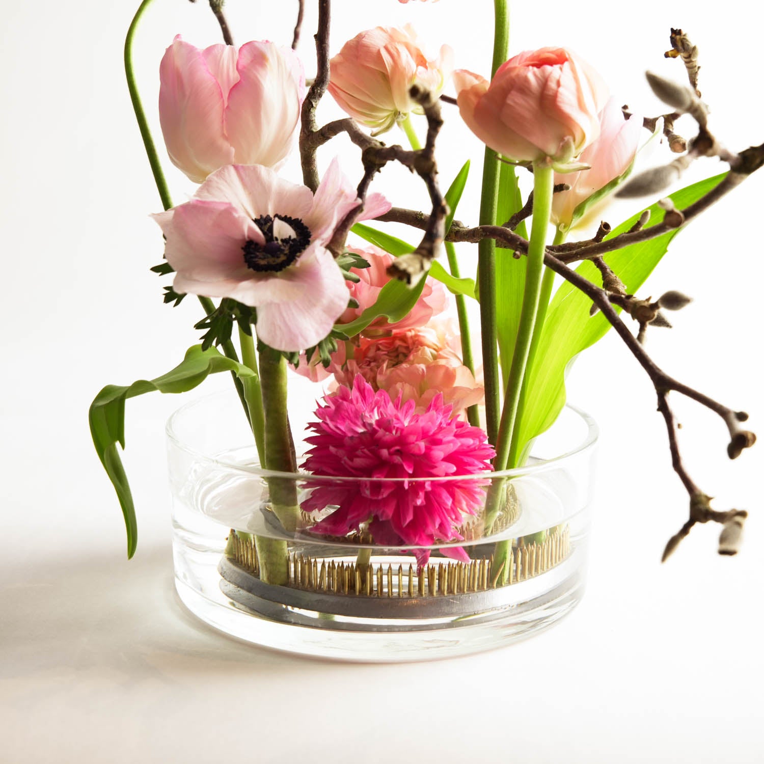 Stunning flower frog arrangement with pink tulips and a small cherry blossom tree branch. The floral arrangement is placed in a clear glass cylinder bowl with water covering the kenzan and hydrating the flower stems. The florist's arrangement is placed in a white setting.