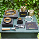 A set of Hanataba flower arrangement tools and accessories arranged on a green wooden surface outdoors, with a leafy green background. The set includes gold and silver metal kenzan rings and brushes, black presentation boxes with gold HANATABA branding, a black packet, a black card, and a small wooden-handled scraper tool.