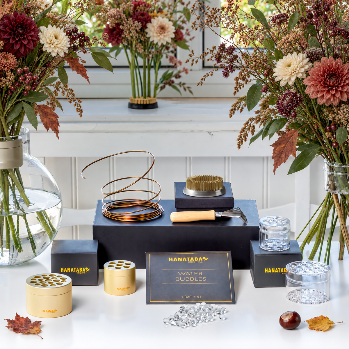 HANATABAS Water Bubbles kit displayed on black risers on a white table, surrounded by autumn floral arrangements and glass vases. The kit includes champagne gold and crystal clear bouquet flower twisters, a kenzan flower frog pin holder ring 70 mm, a wooden-handled kenzan rake, and a sign reading 'HANATABAS WATER BUBBLES'.