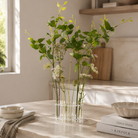 A clear glass vase with a circular base and open top, filled with green stems, small white flowers, and a few pink blossoms, placed on a light stone countertop in a bright kitchen setting.