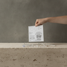 A hand holds a small printed card above a clear acrylic object resting on a rough concrete ledge against a textured gray wall.