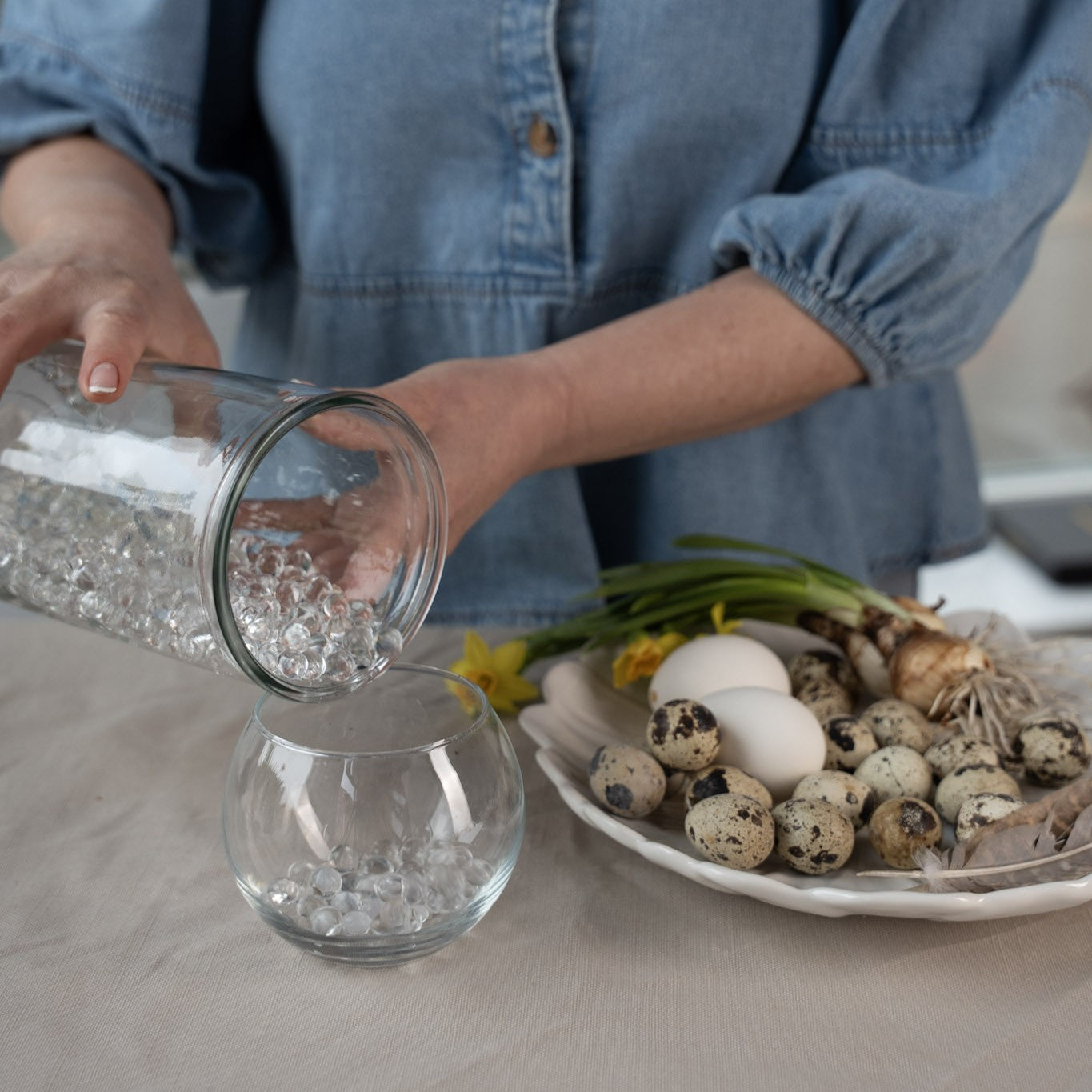 Person pouring hanataba waterbubbles into a glass vase with a plate of eggs and flowers on a table.