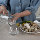 Person pouring hanataba waterbubbles into a glass vase with a plate of eggs and flowers on a table.