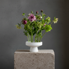 Bouquet of flowers in a white ceramic hanataba ikebana vase on a gray pedestal against a dark gray background