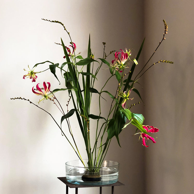 Exotic gloriosa lilies with striking pink and yellow petals, anchored by a 70mm Kenzan ring in a glass dish.
