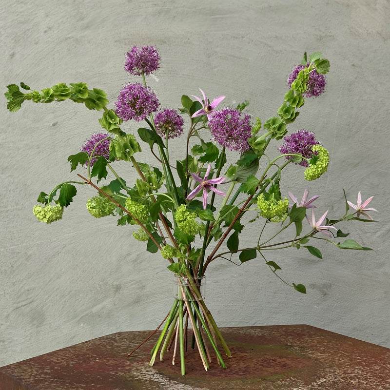 A Hanataba Bouquet Twister secures a vibrant array of purple alliums and delicate pink flowers against a muted wall backdrop.