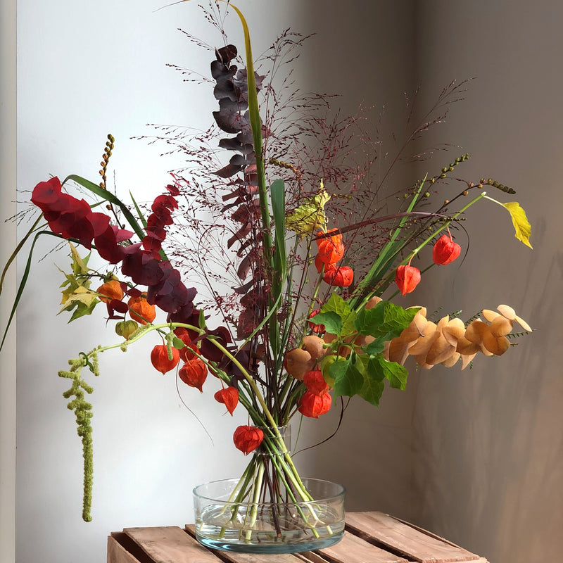 An eclectic Hanataba Bouquet Twister with a vibrant mix of autumnal foliage, including red amaranthus, orange Chinese lanterns, and burgundy smoke bush, in a clear glass bowl.