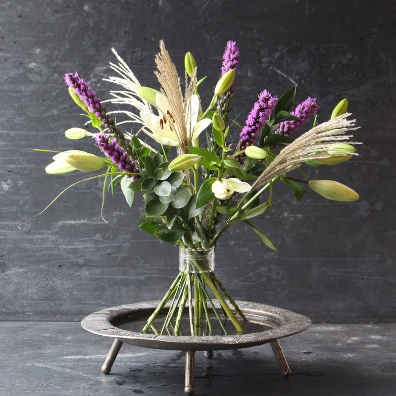 A Hanataba Bouquet Twister Crystal Clear featuring lush greenery, purple flowers, and delicate grasses, arranged in a bouquet twister on a dark background.
