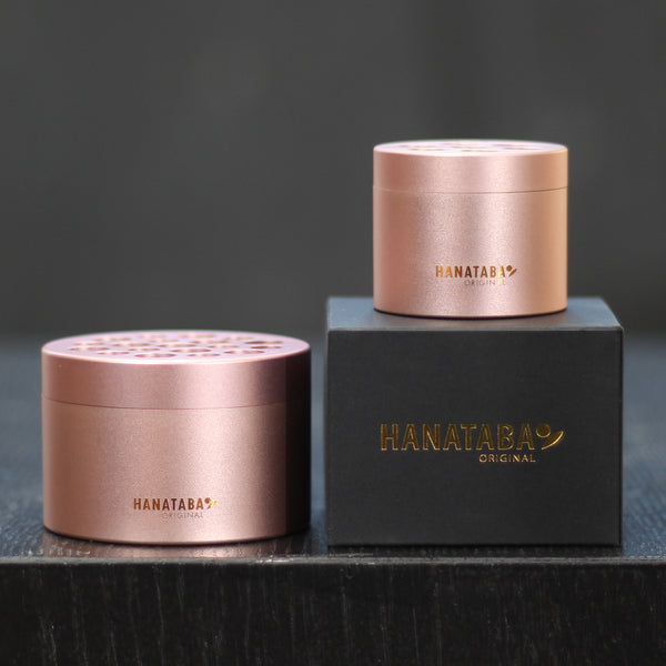 Two Hanataba bouquet twisters in pink champagne, one atop its elegant black box with gold lettering, sit on a dark surface, showcasing the stylish design and functionality of flower arranging tools.