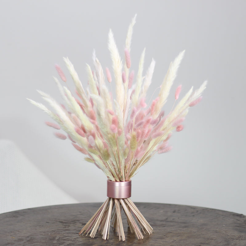 Soft pink and white bunny tail grass is neatly bundled by a Pearly Pink Hanataba bouquet twister, creating a delicate and charming display on a rustic wooden surface.