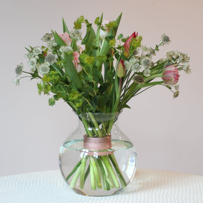 A fresh bouquet of tulips and assorted greenery, secured by a Pearly Pink Champagne Hanataba bouquet twister, sits in a clear, round vase, offering a blend of elegance and springtime charm.