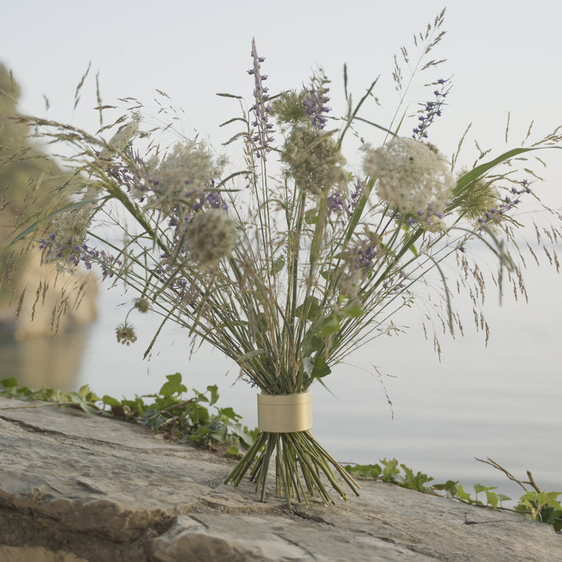 Wildflowers, including purple spikes and white clusters, gathered in a Champagne Gold Hanataba bouquet twister on a rocky ledge overlooking calm waters.