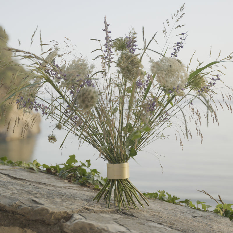 Wildflowers and grasses in a Hanataba Bouquet Twister Champagne Gold, poised on a rock with serene water in the background, conveying natural tranquility.